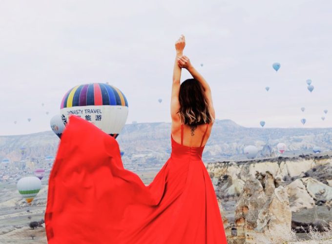 A woman in a flowing red dress standing with Cappadocia’s fairy chimneys and hot air balloon ride in the background.