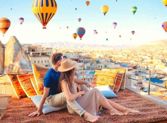 People watching colorful hot air balloons rise into the sky from a terrace in Cappadocia, with a stunning valley view in the background.