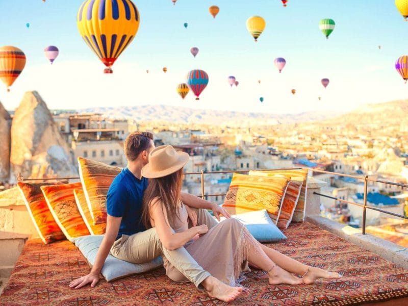 People watching colorful hot air balloons rise into the sky from a terrace in Cappadocia, with a stunning valley view in the background.